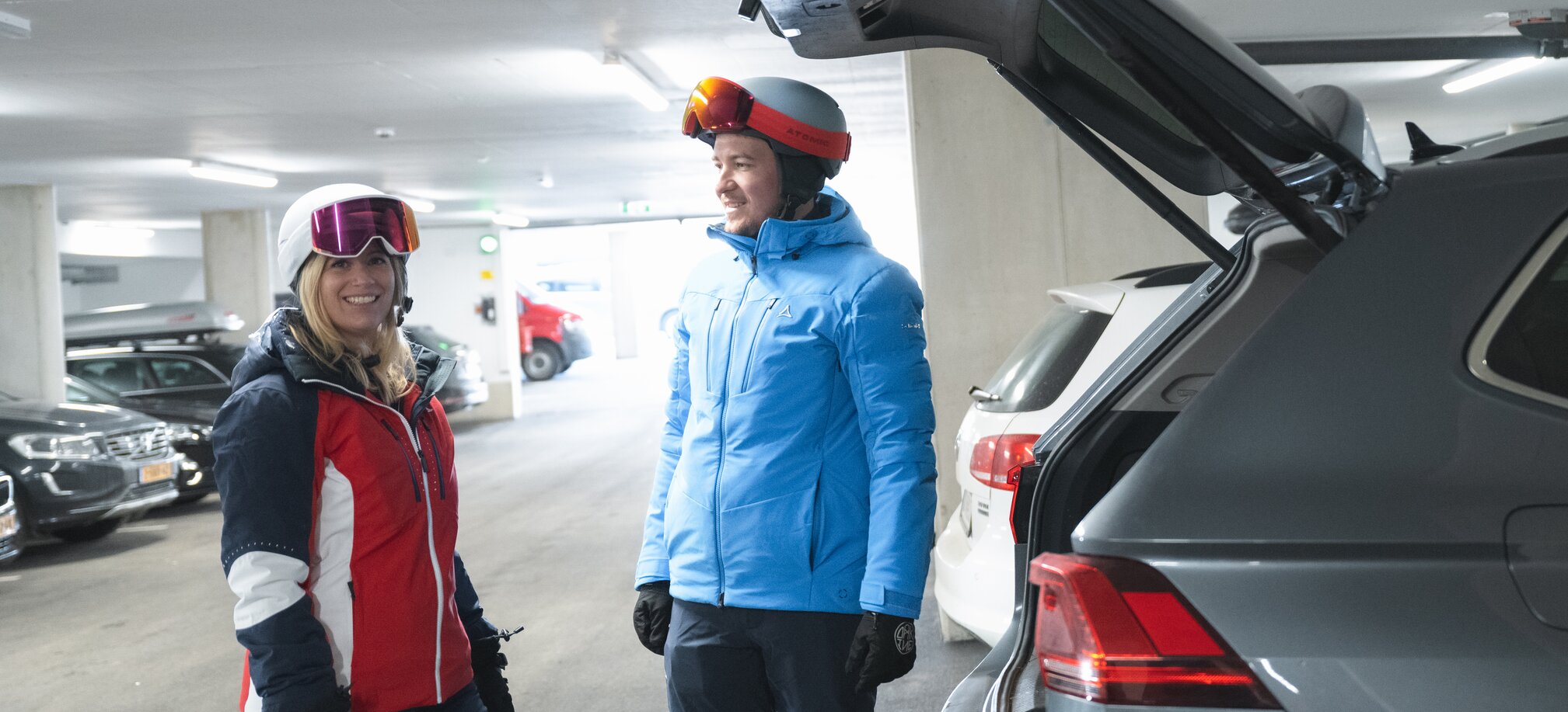 Two people in ski suits and ski helmets stand next to an open boot of a grey car | © Ski amadé