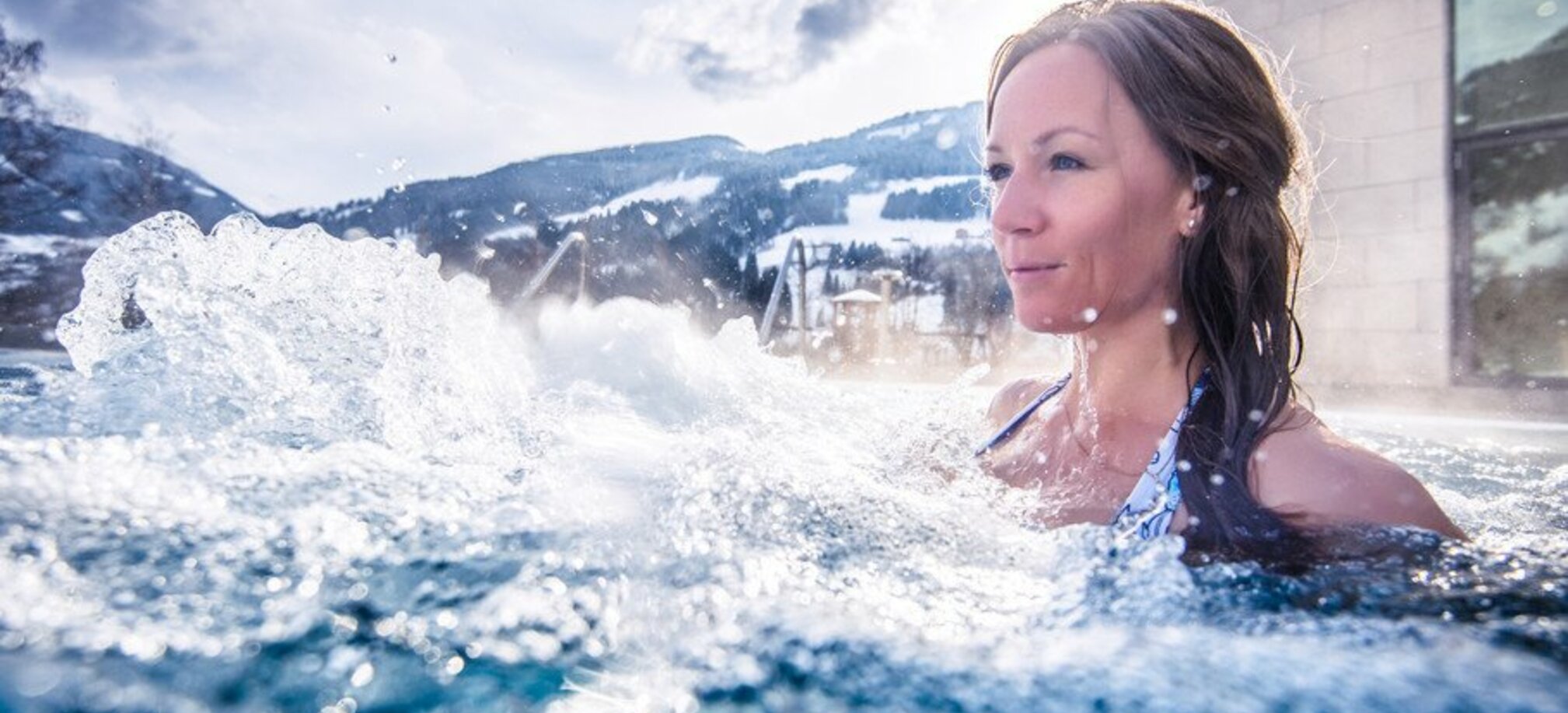 Eine Frau ist in einem Schwimmbecken und vor ihr sprudelt das Wasser und im Hintergrund sind Berge mit Skipisten zu sehen