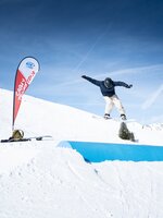 Snowboarder jumps over blue tube feature at Cash4Tricks with clear blue sky, red event flag and gear placed on the snow nearby.