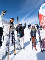 Four young freeskiers cheer in front of a Cash4Tricks flag, holding skis in the snow with blue sky and mountains behind them.