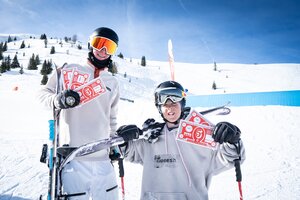 Two young freeskiers hold red Cash4Tricks vouchers, posing in snowpark scenery with skis and blue park tube behind them.