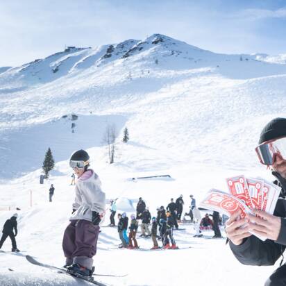 A judge kneels in snow holding red Cash4Tricks vouchers, spectators under bright alpine sun.