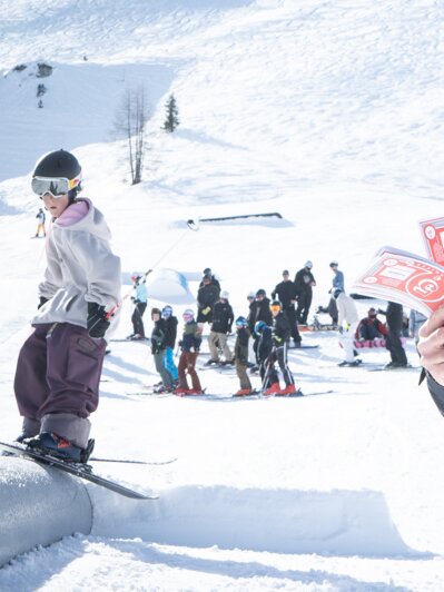 Ein Judge kniet im Schnee mit roten Cash4Tricks-Gutscheinen, viele Zuschauer bei strahlendem Sonnenschein.