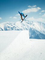 Snowboarder in blauem Hoodie zeigt Grab-Trick über Kicker im Snowpark mit verschneiten Alpen und Sonnenschein.