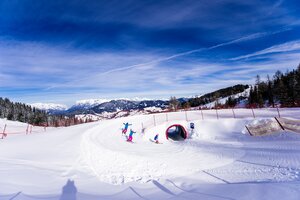 Drei Kinder auf Skiern fahren durch einen Schneetunnel auf der Funslope bei Sonne und mit Bergpanorama im Rücken.