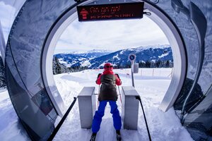 Kind mit Helm und Stöcken im Starttor einer Kinderrennstrecke mit Blick auf verschneite Hänge und Berge.