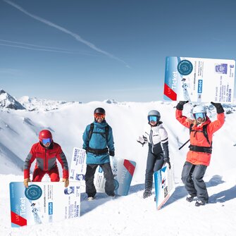 Four winter sports enthusiasts pose with giant ski passes in the snow before alpine mountain scenery.