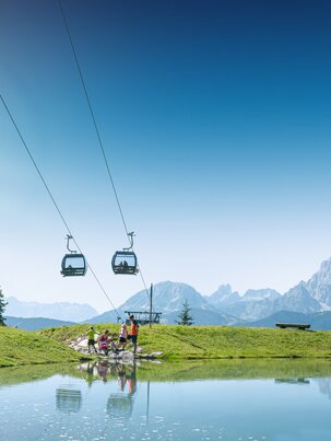 Family by the shore of a small mountain pond beneath a lift, with green meadows and Alps behind