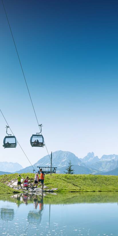 Family by the shore of a small mountain pond beneath a lift, with green meadows and Alps behind