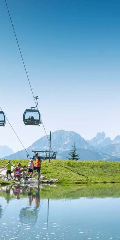 Family by the shore of a small mountain pond beneath a lift, with green meadows and Alps behind