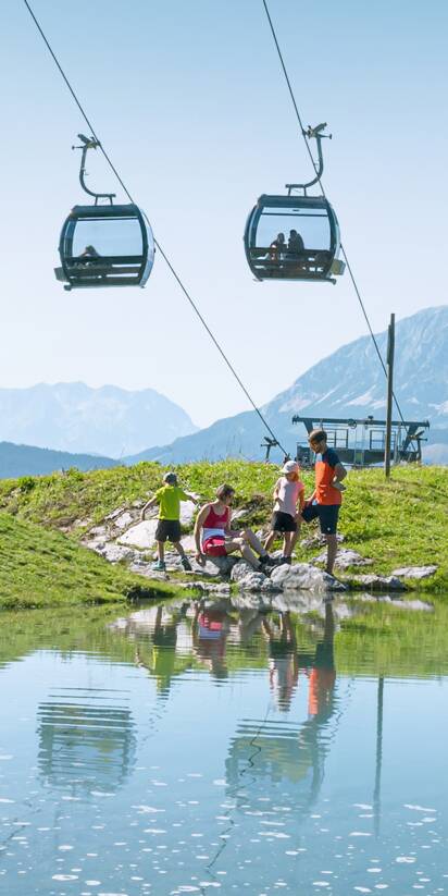 Family by the shore of a small mountain pond beneath a lift, with green meadows and Alps behind