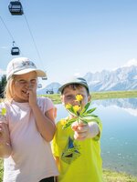 Zwei Kinder mit gepflückten Blumen stehen am Bergsee in Ski amadé, im Hintergrund die markanten Dachstein-Berge.