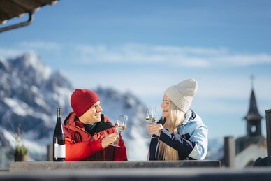Man and woman toast with white wine, snowy mountains and church tower visible in background | © Ski amadé