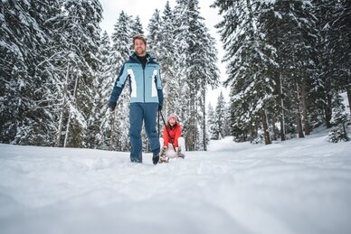 Ein Mann zieht eine lachende Frau auf einem Schlitten durch einen verschneiten Waldweg. | © Ski amadé