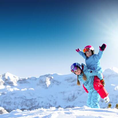 Two children in colorful ski outfits laugh and play in the snow before snowy peaks