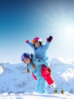 Two children in colorful ski outfits laugh and play in the snow before snowy peaks