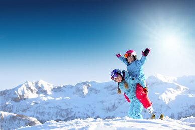 Zwei Kinder in bunter Skikleidung spielen lachend im Schnee vor verschneiten Bergen