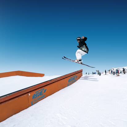 Skier jumps over rail element in snow park, other winter sports fans watching in the background