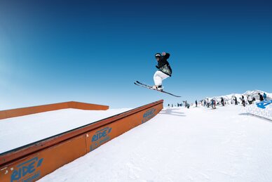 Skier jumps over rail element in snow park, other winter sports fans watching in the background