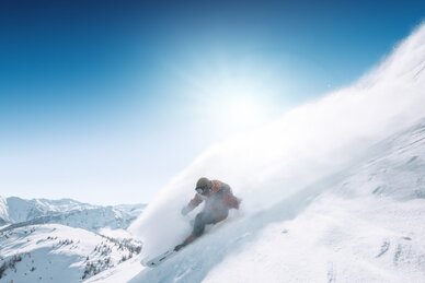 Snowboarder fährt im Tiefschnee eine steile Hangflanke hinab, Pulverschnee spritzt auf