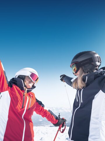 Two female skiers face each other with arms wide open, smiling in the snow and sunshine