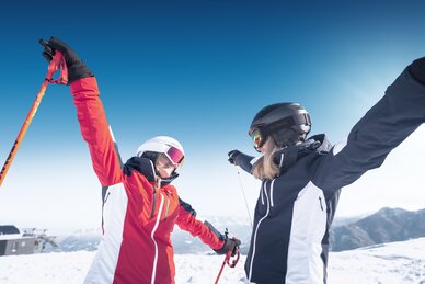 Two female skiers face each other with arms wide open, smiling in the snow and sunshine