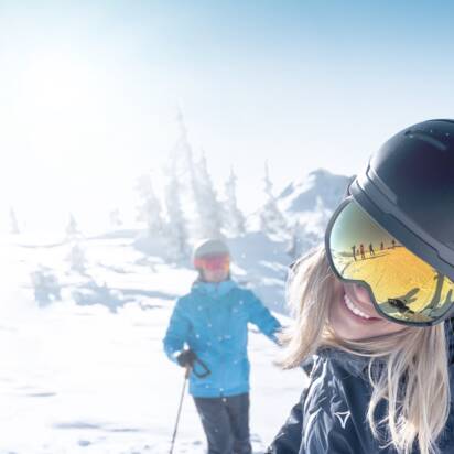 Woman with helmet and ski goggles smiles at camera, two skiers in snow behind her