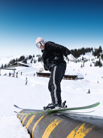 Young female skier balances on yellow-black rail in snow park on a sunny winter day