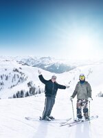 Zwei Skifahrer stehen auf einem sonnigen Hang mit weitem Blick auf schneebedeckte Berge