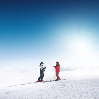 Two skiers stand on freshly groomed slope in Ski amadé overlooking a foggy valley in bright sunlight