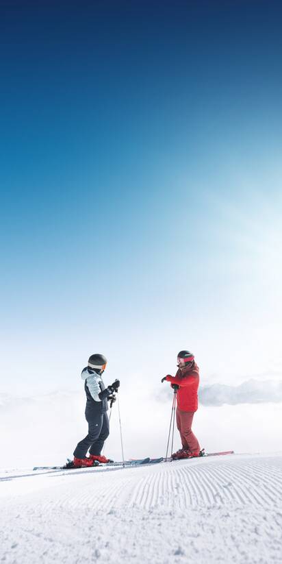 Two skiers stand on freshly groomed slope in Ski amadé overlooking a foggy valley in bright sunlight