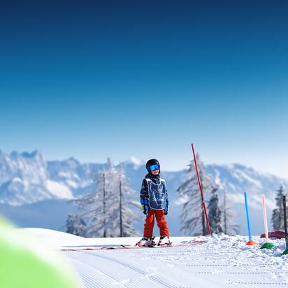 Child wearing helmet and vest stands on sunny training slope with skis in front of snowy mountains