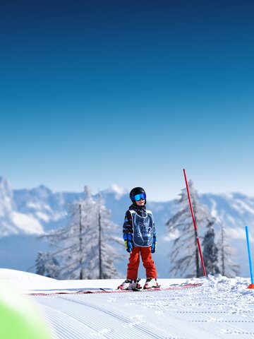 Kind mit Helm und Weste steht auf sonniger Übungspiste mit Skiern vor verschneiten Bergen