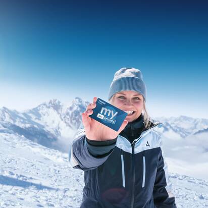 Woman in ski jacket holds my Ski amadé Friends Card in front of snowy Alps under bright sunshine