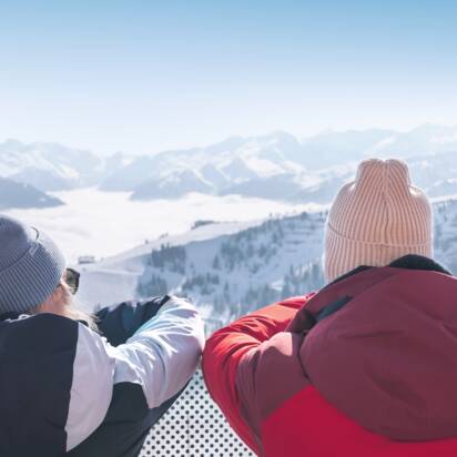 Two people in winter clothes stand on terrace overlooking snowy mountains with skis in the foreground