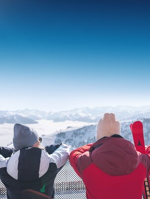 Zwei Personen in Winterkleidung stehen auf Terrasse und schauen auf verschneite Berge mit Skiern im Vordergrund