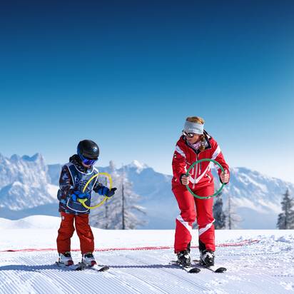 Skilehrerin und Kind mit bunten Ringen üben auf sonniger Piste im Schnee bei Best Learn2Ski amadé Kurs