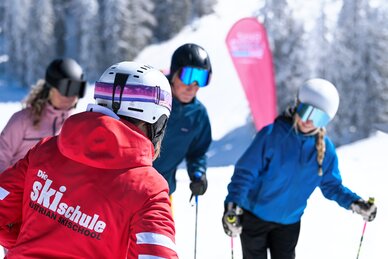 Ski instructor in red jacket teaches group of skiers with helmets and goggles on sunny slope in Ski amadé