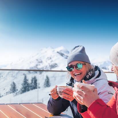 Two people in ski jackets drink hot beverages on terrace with view of snowy mountains in bright sunshine
