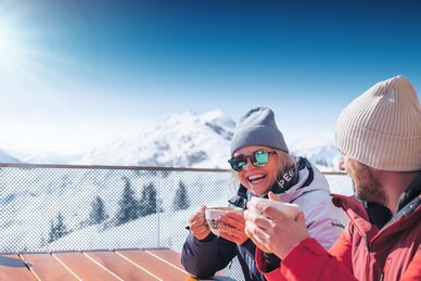 Two people in ski jackets drink hot beverages on terrace with view of snowy mountains in bright sunshine