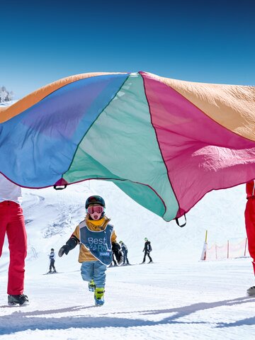 Kinder in Best Learn2Ski-Westen spielen unter buntem Schwungtuch auf verschneiter Piste mit Skilehrern im Hintergrund