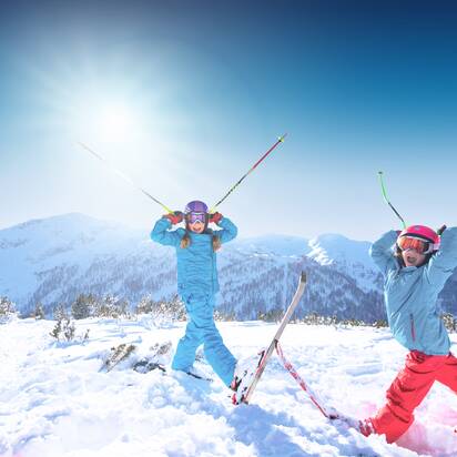 Two children are standing in deep snow, holding their ski poles to their helmets so that they look like horns, with their mouths wide open