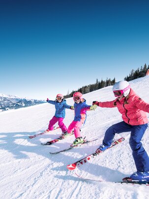 Three children hold hands and ski down the piste side by side, their ski tips touching and they laugh as they do so