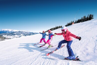 Three children hold hands and ski down the piste side by side, their ski tips touching and they laugh as they do so
