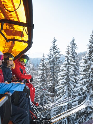 Four skiers wearing helmets and goggles sit on a chairlift with an orange cover above snow-covered fir trees. | © Ski amadé