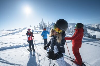 Four skiers enjoy the sun on a snow-covered piste. A woman in the foreground laughs into the camera. | © Ski amadé
