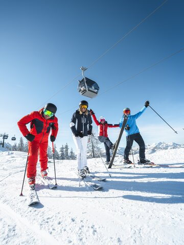 Skiers rejoice and stand on the piste with a cable car hovering above them in the bright blue sky. | © Ski amadé