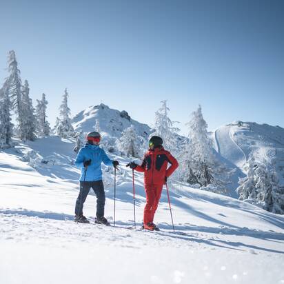 Two men in red and blue ski suits chatting on the piste, with beautiful snowy scenery in the background | © Ski amadé