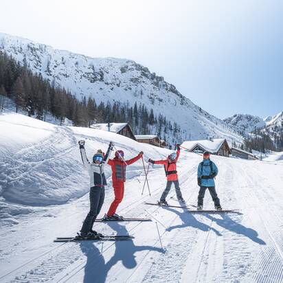 Four winter athletes cheer in front of alpine huts in snowy valley, surrounded by mountains, sun and blue sky.