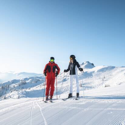 Two skiers stand side by side on freshly groomed slope with sunshine, chairlift and Alpine view